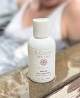 A bottle of 5 Circle Skin Care Naked Oil Cleanser sits on a stone surface with a blurred person in a white top in the background.