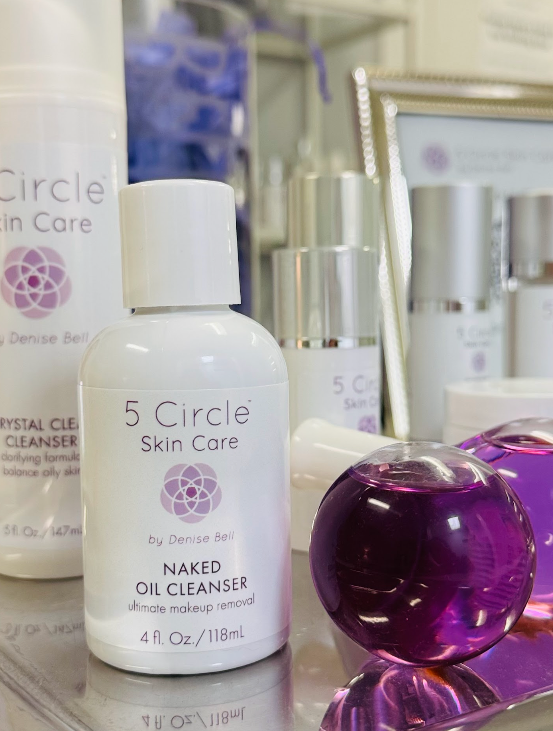 A bottle of 5 Circle Skin Care Naked Oil Cleanser in the nude color, shown alongside other skincare products in a reflective, premium setting.
